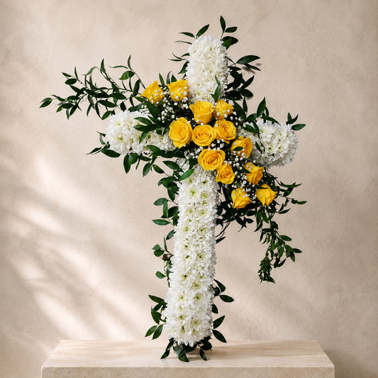 A floral cross arrangement featuring white hydrangeas, white carnations, yellow roses, and green foliage, displayed on a light stone pedestal against a textured beige wall.