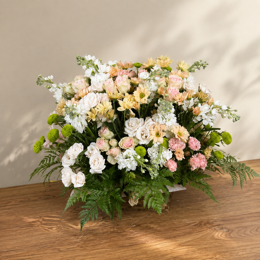 Gentle Farewell pastel floral basket with white and peach flowers, lush greenery and a woven base.