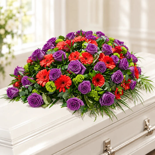 A vibrant funeral flower arrangement featuring purple roses, red gerbera daisies, and green foliage, displayed on a white casket.
