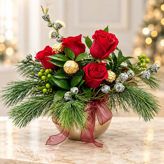 Christmas roses with chocolates and pine greenery in a gold vase with a burgundy bow on a marble table.