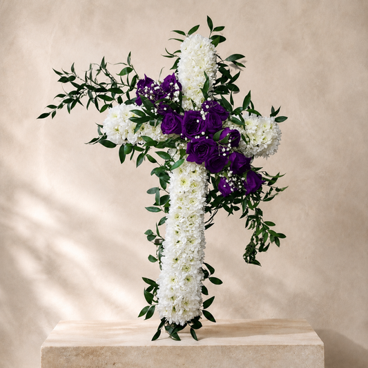 A large decorative floral cross made of white flowers and greenery, with prominent purple roses in the center, displayed on a stone pedestal against a warm beige wall.