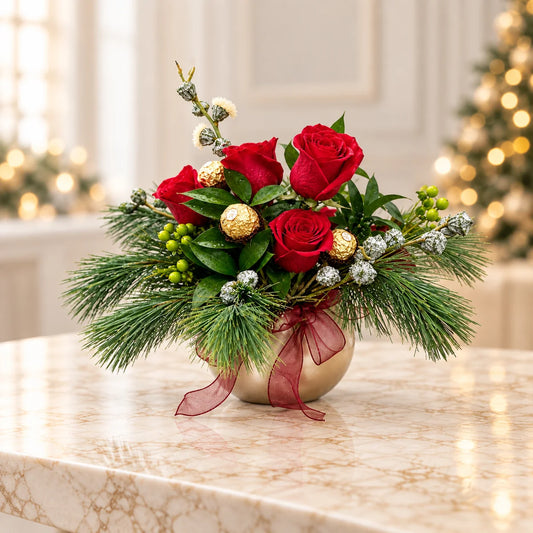 Golden Christmas flower arrangement in a gold vase on marble, with red roses, chocolates and warm holiday lights behind.