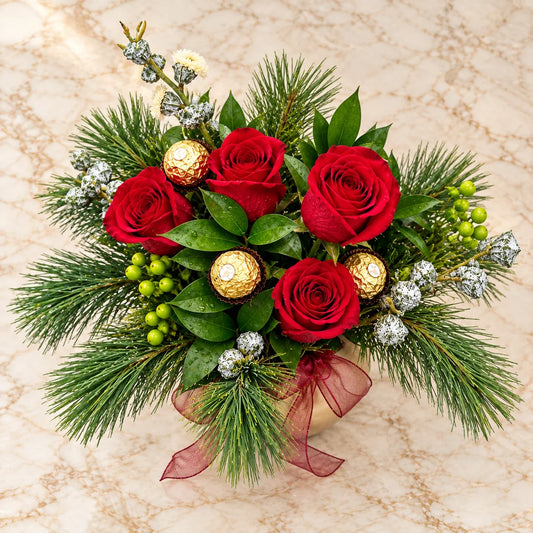 Overhead view of red roses, gold-wrapped chocolates, green berries and pine foliage in the Golden Christmas arrangement.