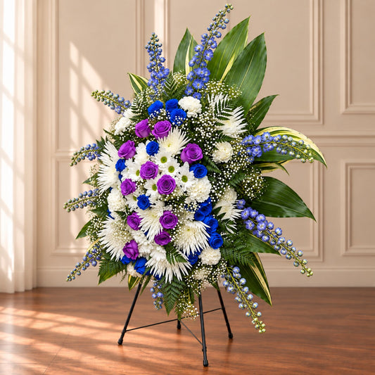 Purple roses, blue delphinium and white funeral flowers in a standing tribute by Amarilis Flowers